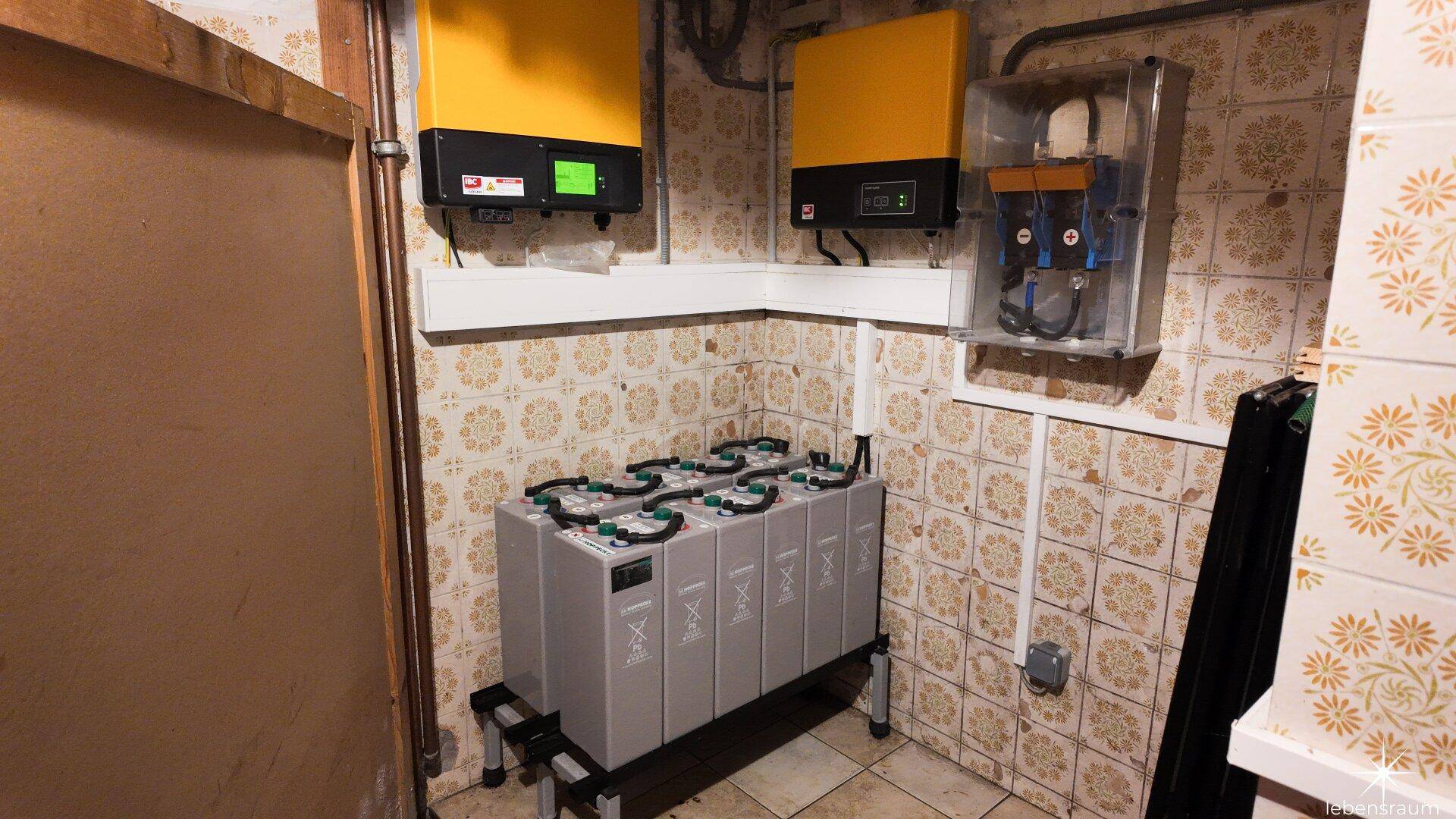 Technikraum mit Batterien und elektrischen Installationen an gefliesten Wänden.