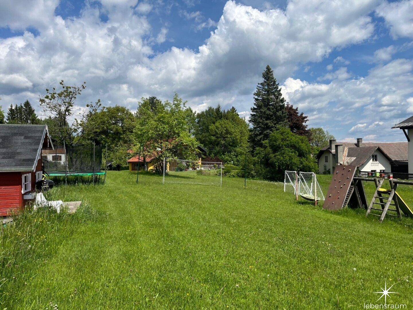 Großer Rasenbereich mit Trampolin, Spielplatz und Häusern im Hintergrund, umgeben von Natur.