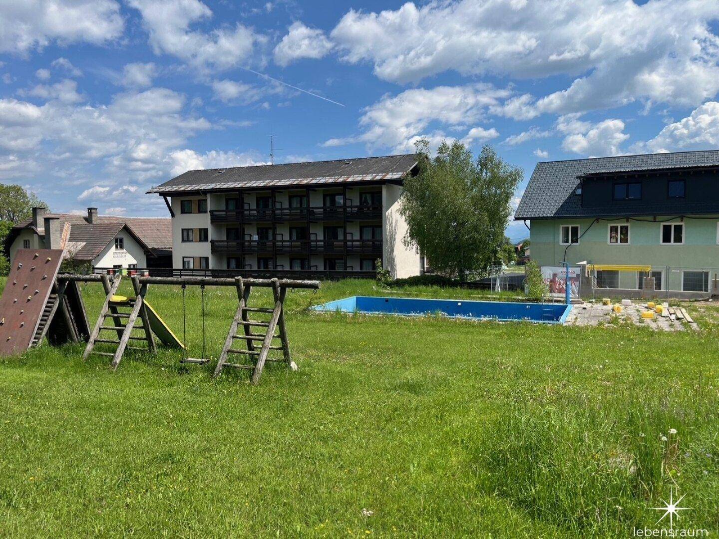 Großer Gartenbereich mit Spielplatz und Pool, umgeben von Mehrfamilienhäusern unter blauem Himmel.