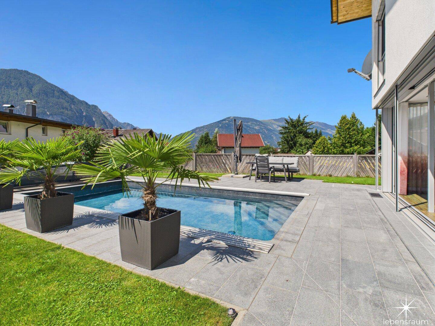 Großer Poolbereich mit Terrasse, Rasenfläche und Bergblick an einem sonnigen Tag.