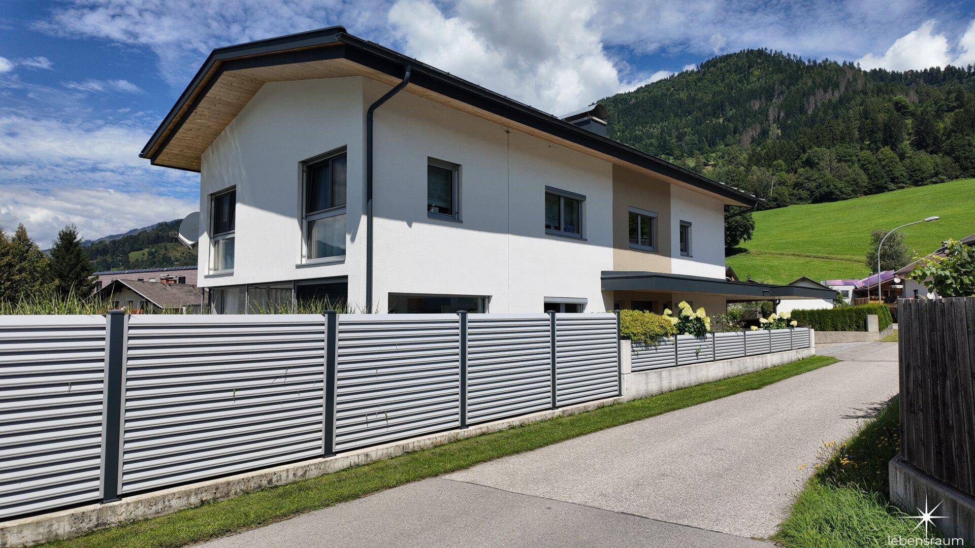 Moderne Hausfassade mit Zaun und grüner Umgebung in den Bergen.