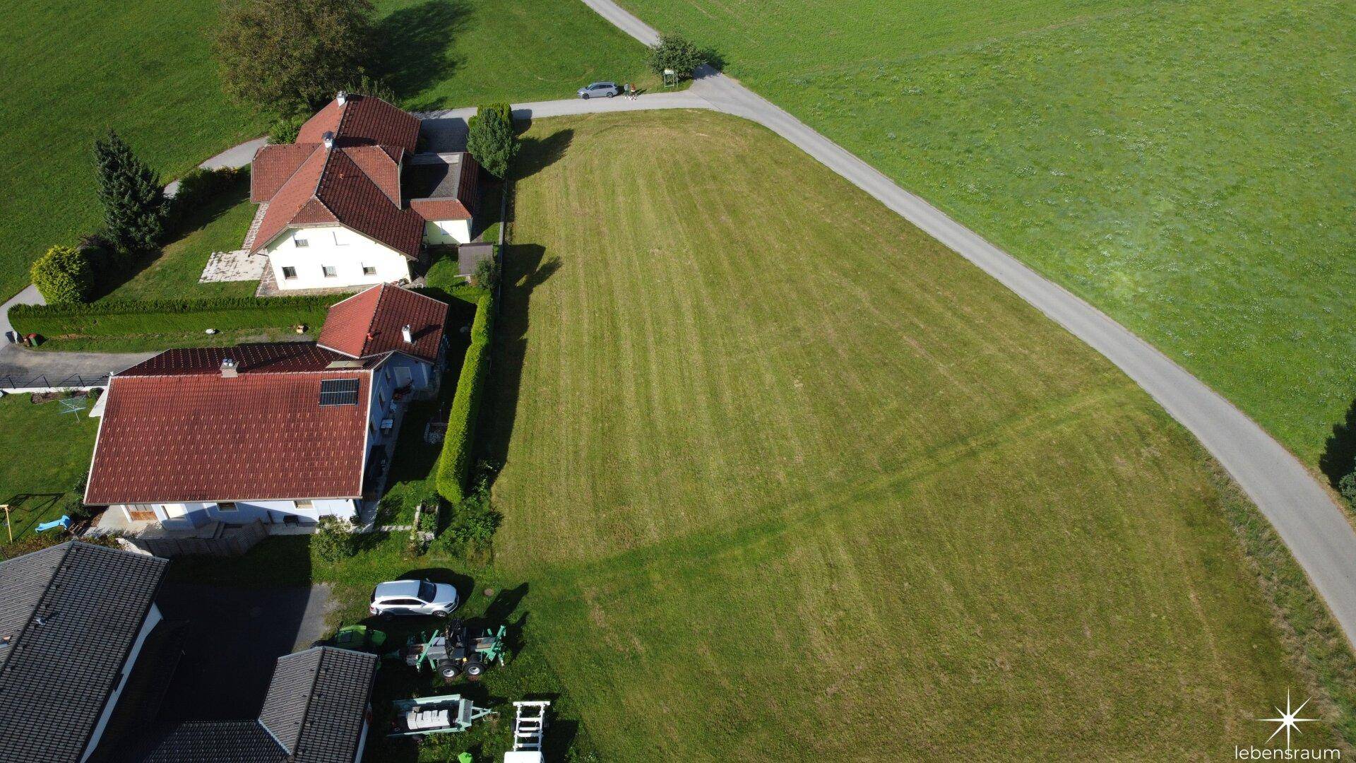 Luftaufnahme eines weitläufigen grünen Grundstücks, angrenzend an Wohnhäuser mit roten Dächern und eine Landstraße.