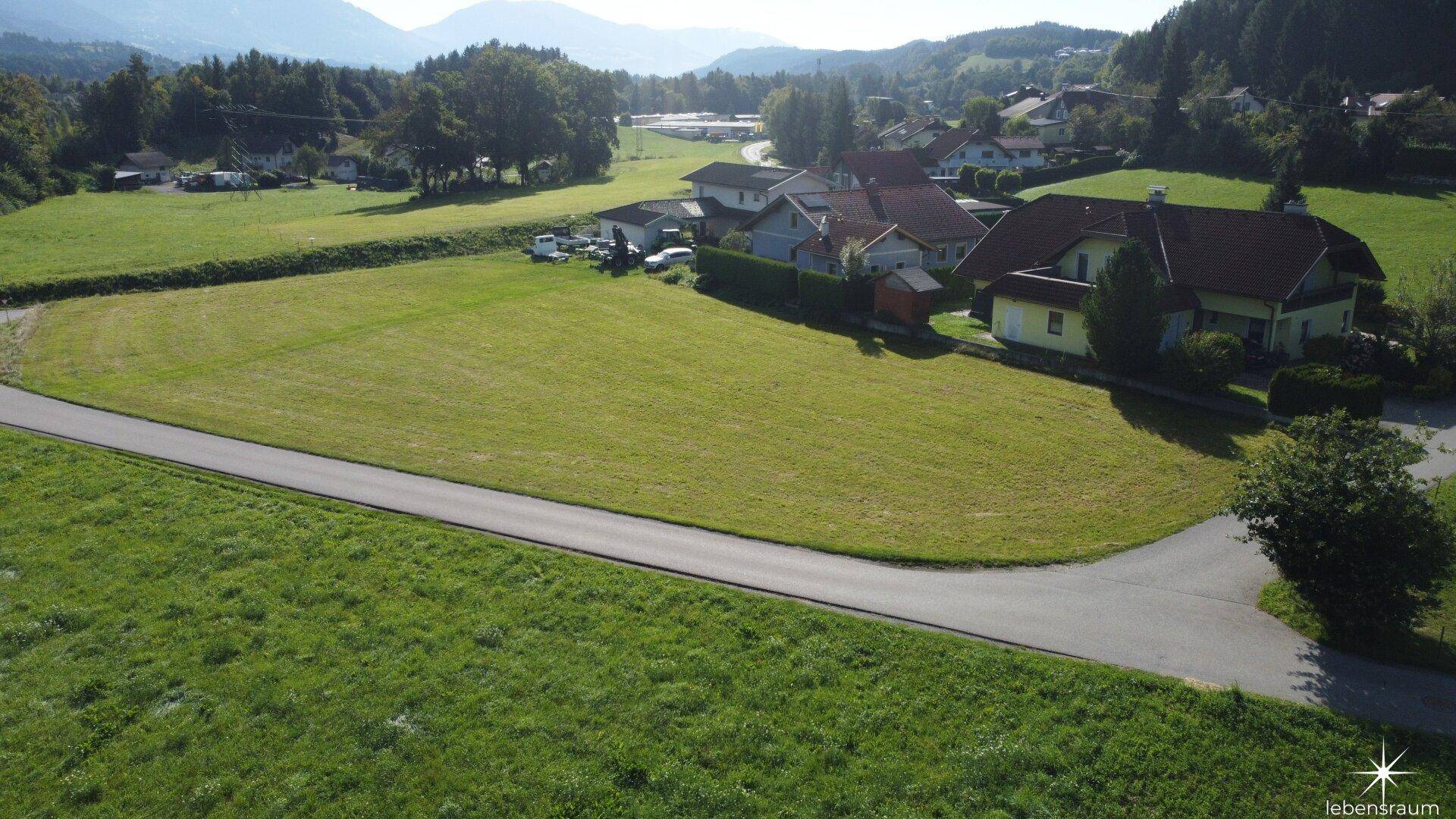 Panoramische Luftaufnahme eines weitläufigen grünen Grundstücks mit ländlicher Bebauung und Bergsilhouette im Hintergrund.