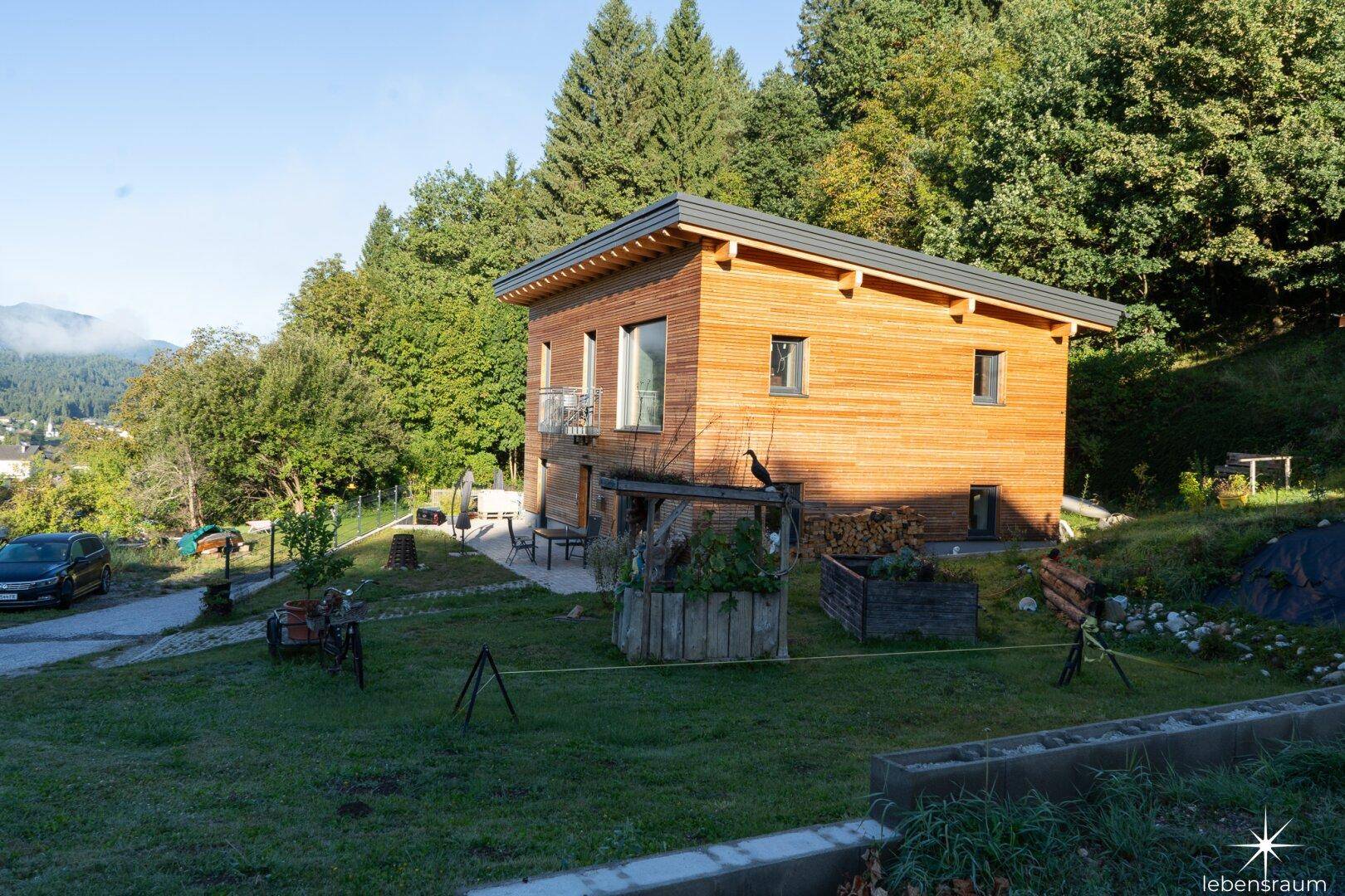 Modernes Holzhaus mit Gartenbereich und Hochbeeten in sonniger, grüner Umgebung.
