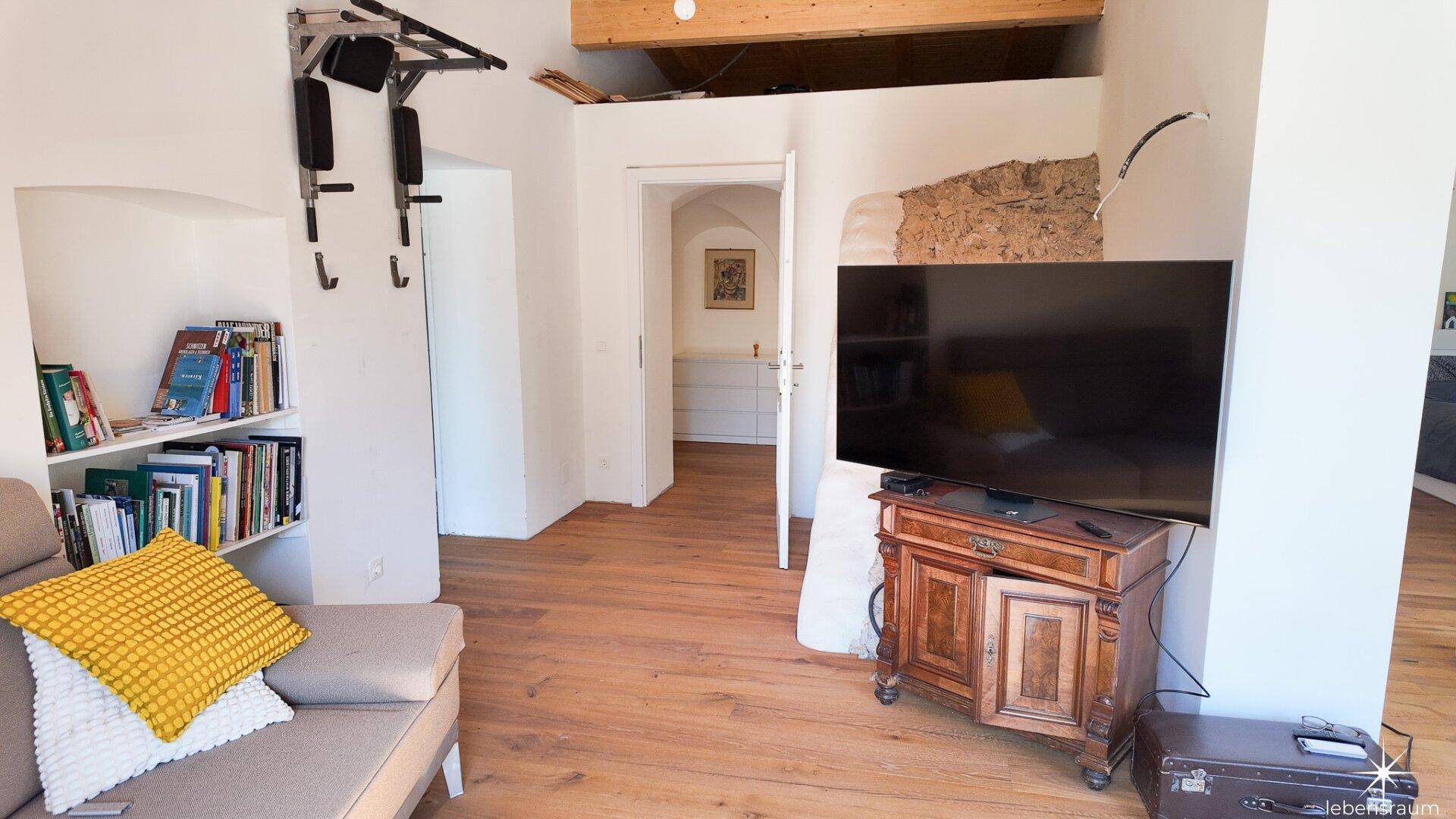 Wohnbereich mit Sofa, Bücherregal und Fernseher, führt zu einem weiteren Raum.