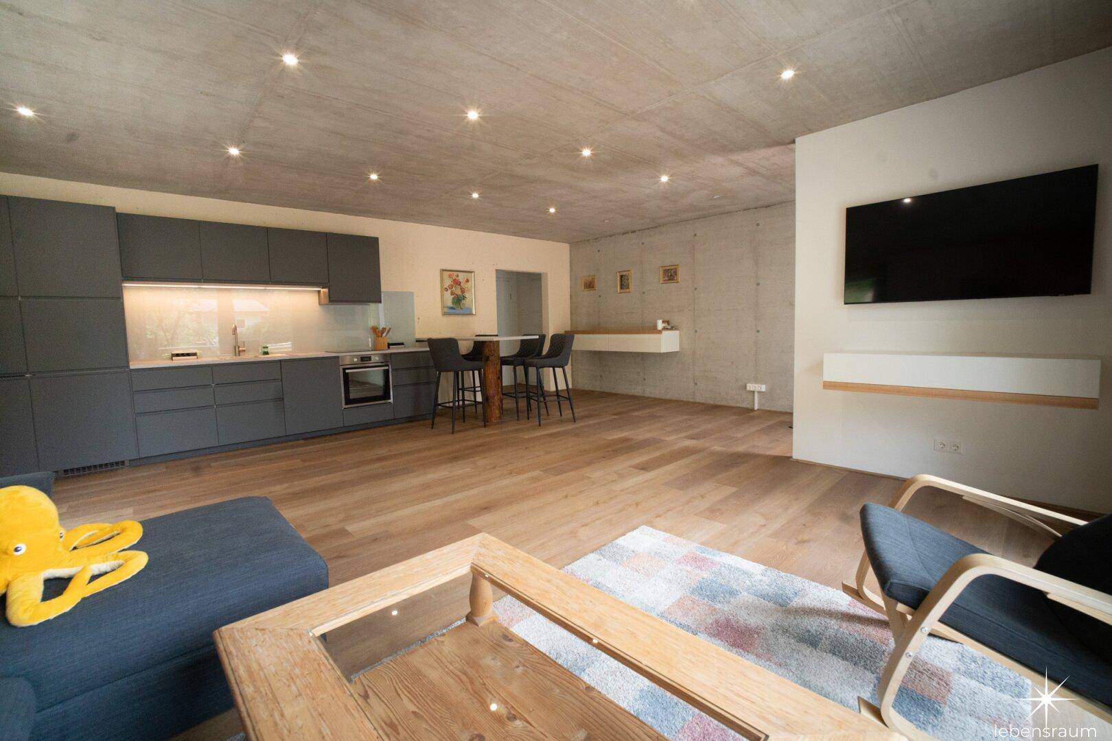 Moderner Wohn- und Essbereich mit offener Küche, Holzboden und Sichtbetondecke.