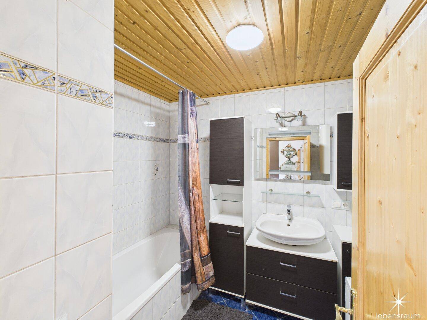 Badezimmer mit Badewanne, Waschbecken und Holzdecke, gefliest in Weiß und Blau, sauber und gepflegt.