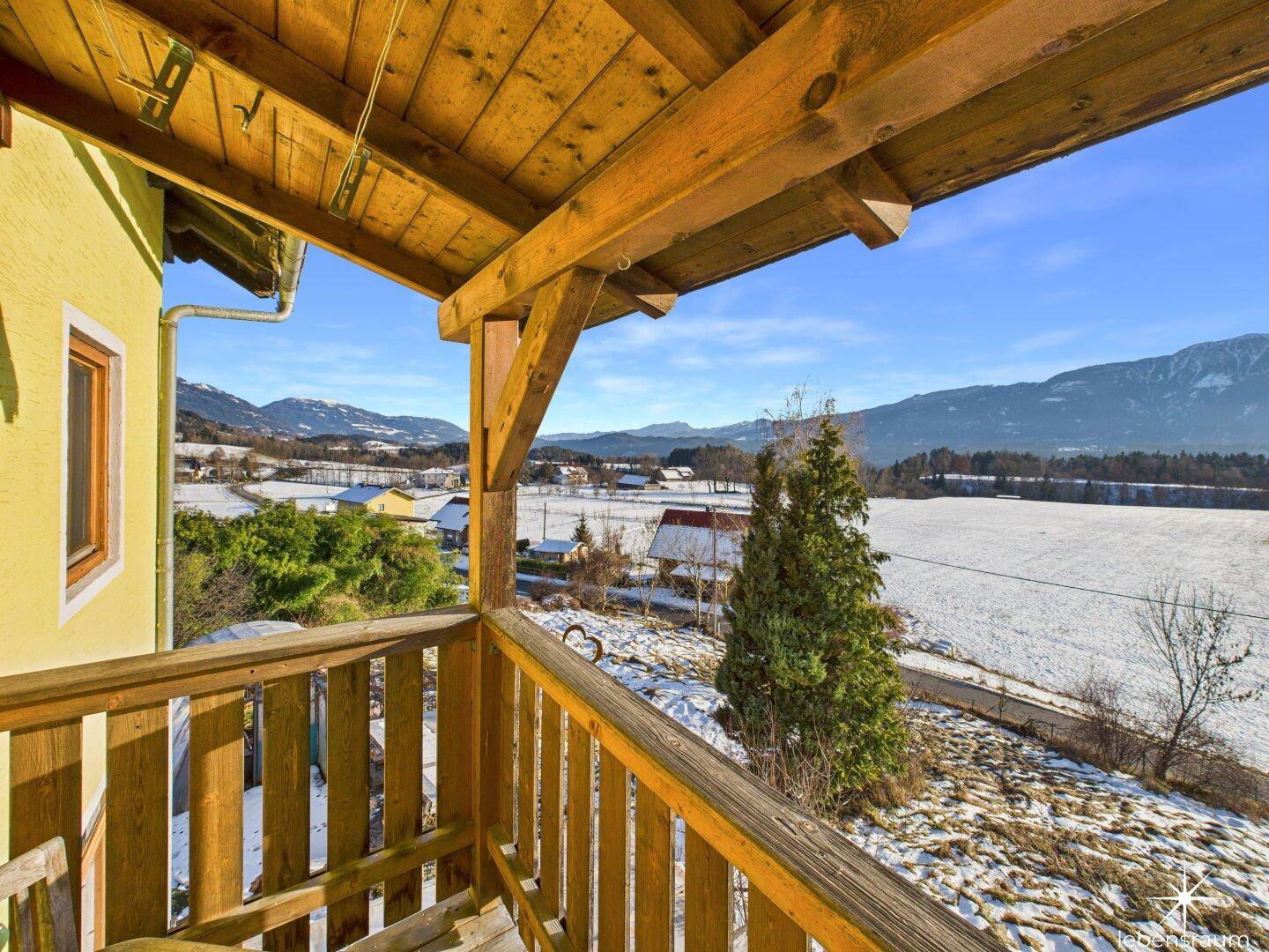 Rustikaler Holzbalkon mit weitem Blick über die verschneite Landschaft und Berge unter blauem Himmel.