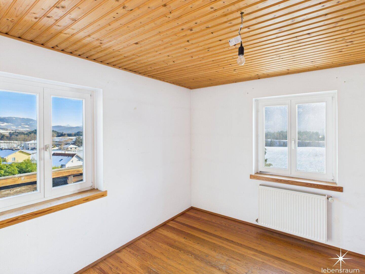 Helles Zimmer mit Holzboden, Holzdecke und zwei Fenstern mit Ausblick auf die Natur.