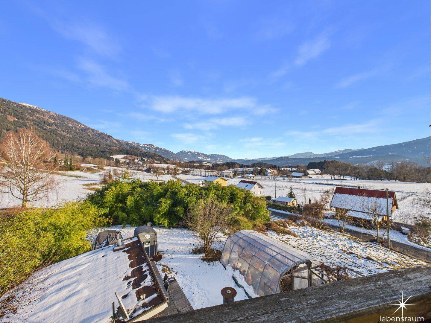 Weitläufiger Blick über die verschneite Landschaft mit Bergen und grünen Büschen unter blauem Himmel.
