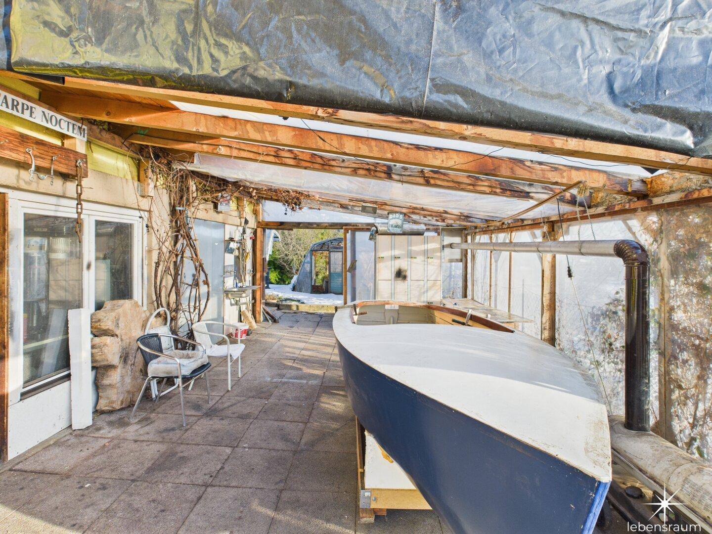 Überdachter Bereich, möglicherweise ein Wintergarten, mit einem Boot und Sitzgelegenheiten, rustikal gestaltet.