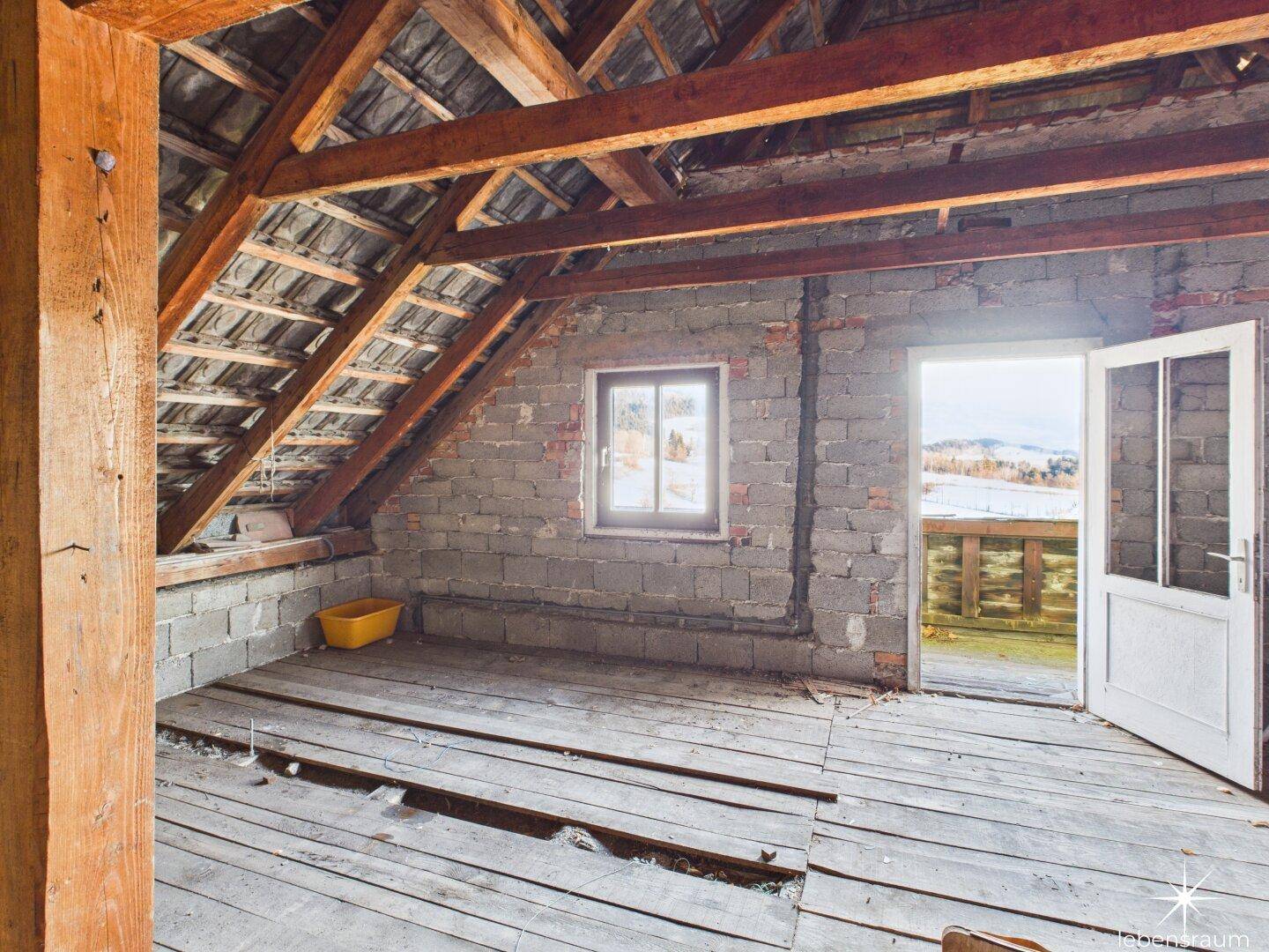 Unausgebauter Dachboden mit Holzbalken, Ziegelwänden und Blick nach draußen, bietet Ausbaupotenzial.