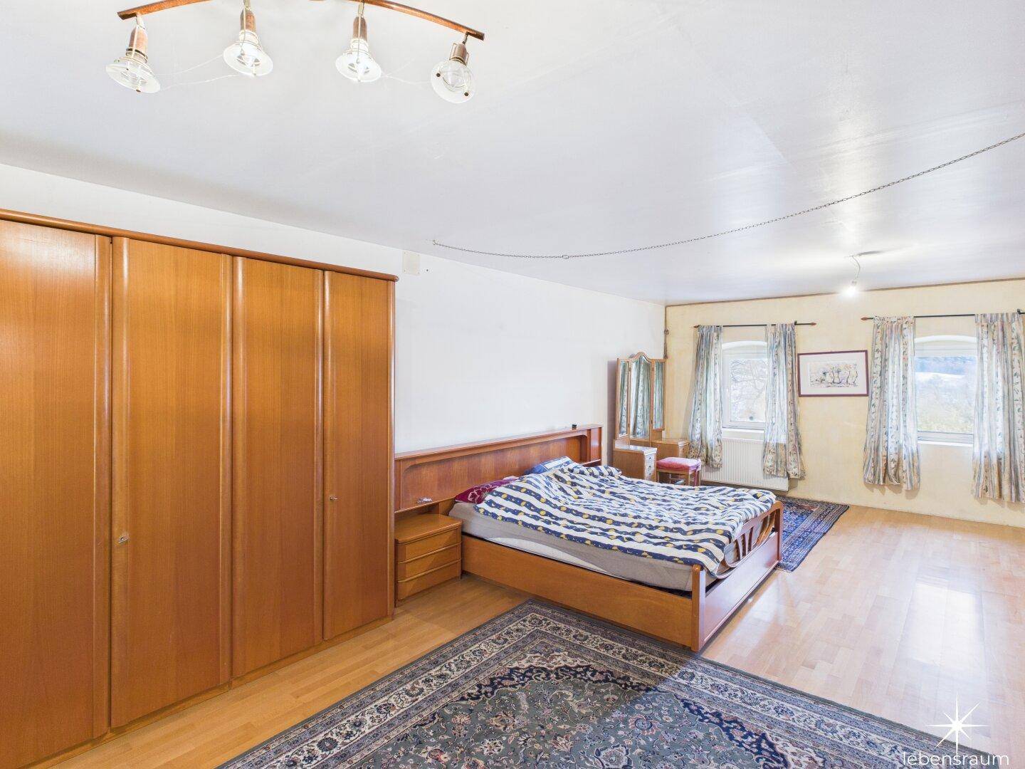 Schlafzimmer mit großem Bett, Holzschrank und Fenstern mit Blick ins Grüne, gemütlich und hell.