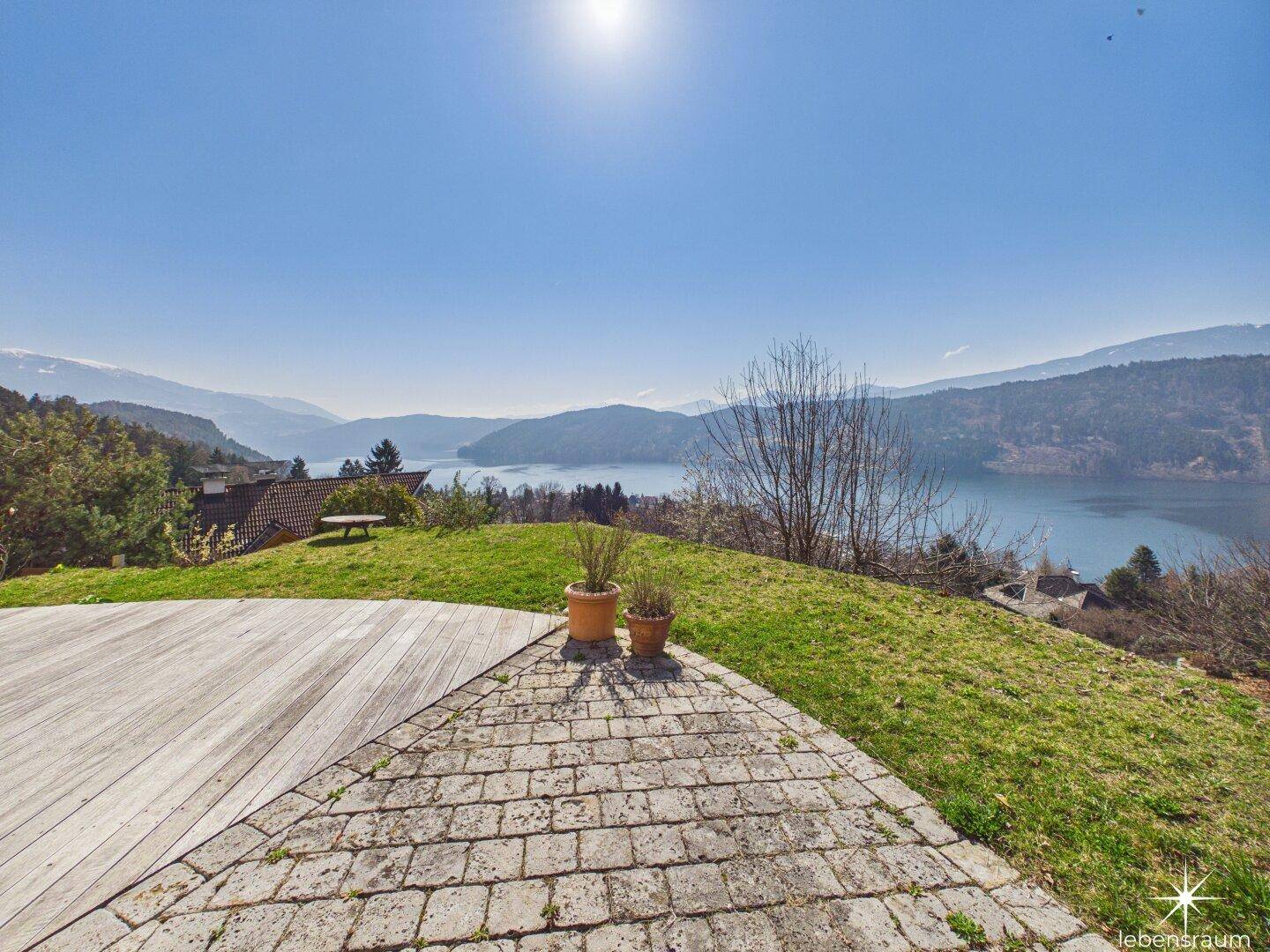 Terrasse mit Holzdeck und Steinplatten, umgeben von Grünflächen und Panoramablick auf den See.