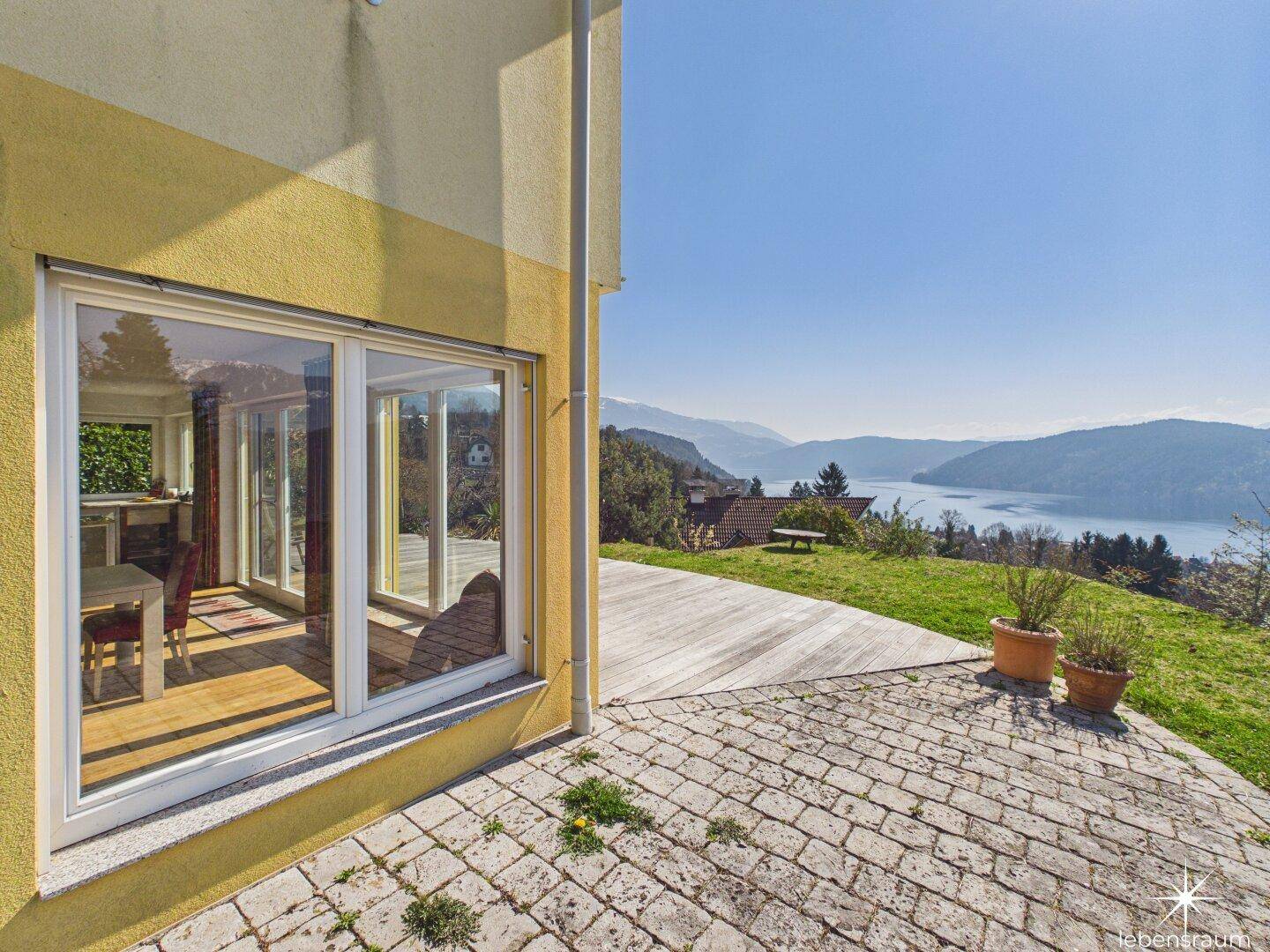 Terrasse mit Steinplatten und Holzdeck, angrenzend an das Haus mit großen Fenstern und Seeblick.