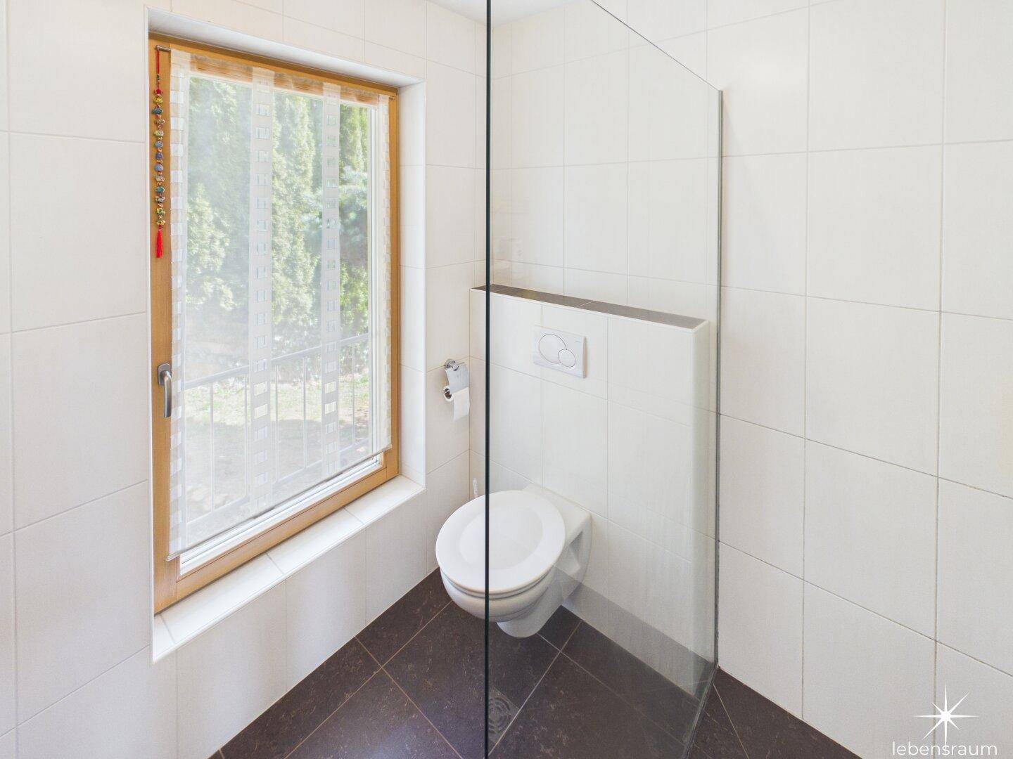 Modernes WC mit wandhängender Toilette, Fliesen und Fenster mit Blick ins Grüne.