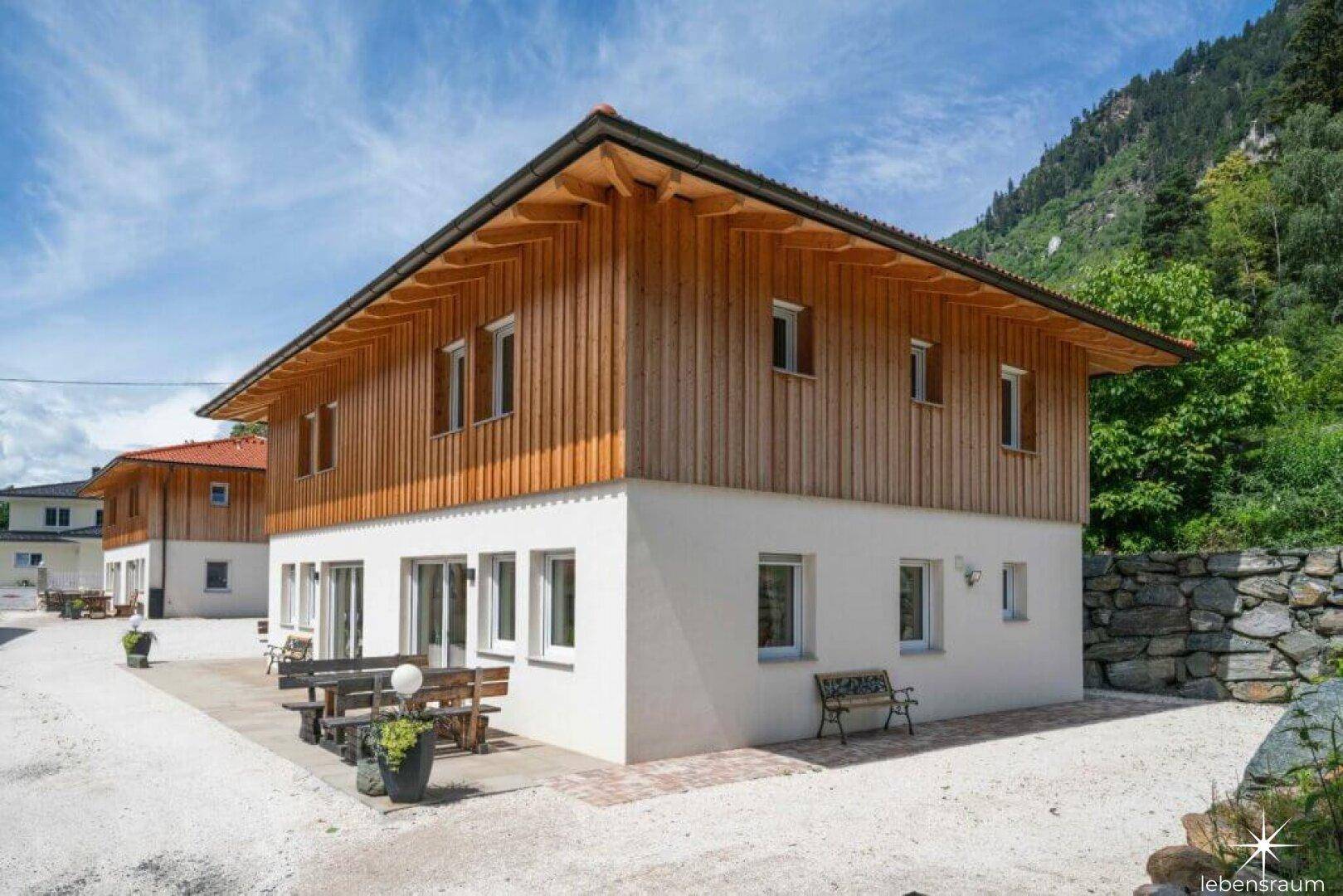 Seitenansicht des Hauses mit Holzverkleidung und gepflasterter Terrasse vor bergiger Kulisse.