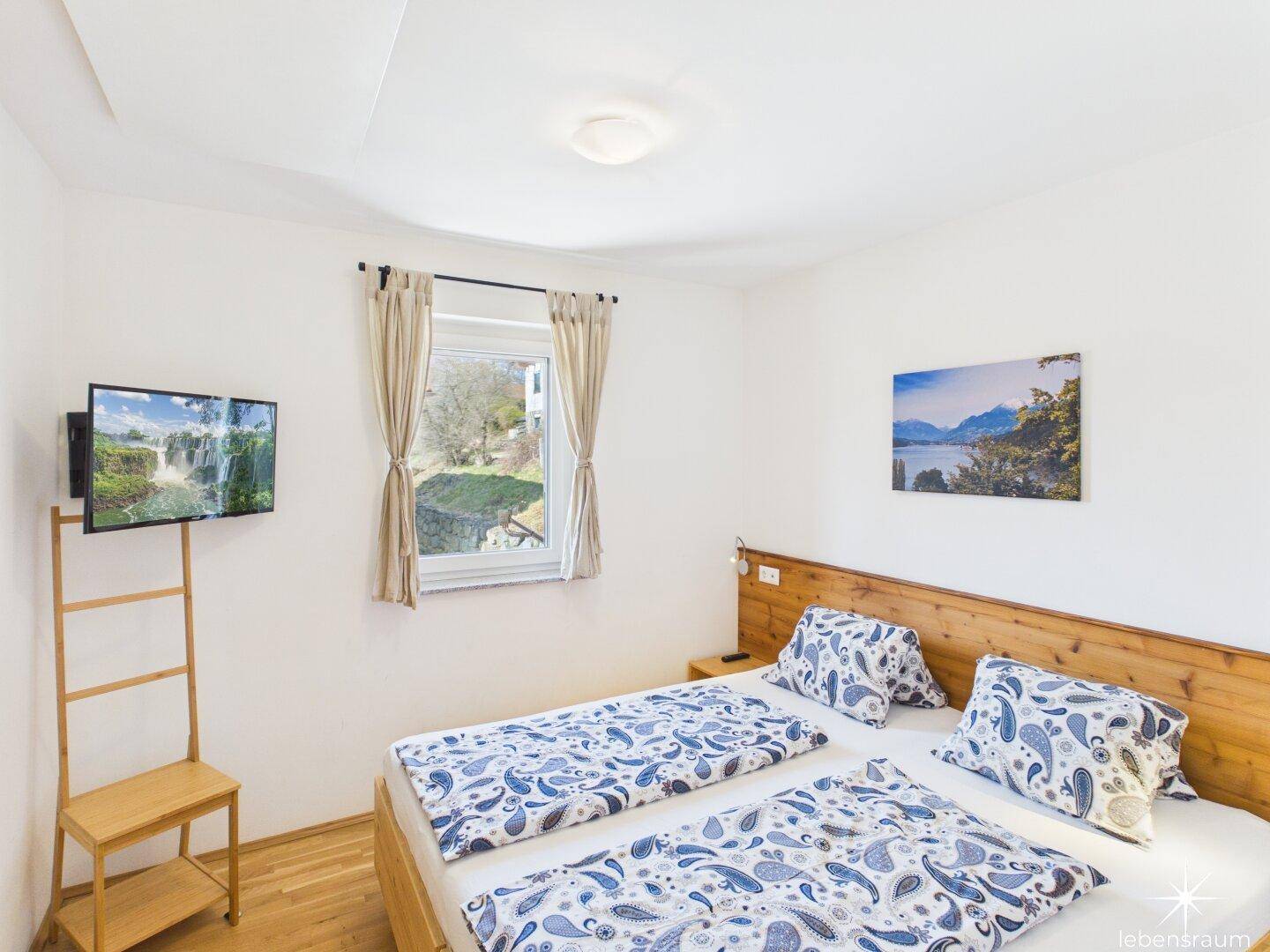 Helles Schlafzimmer mit Doppelbett, Holzkopfteil und Fenster mit Blick ins Grüne.