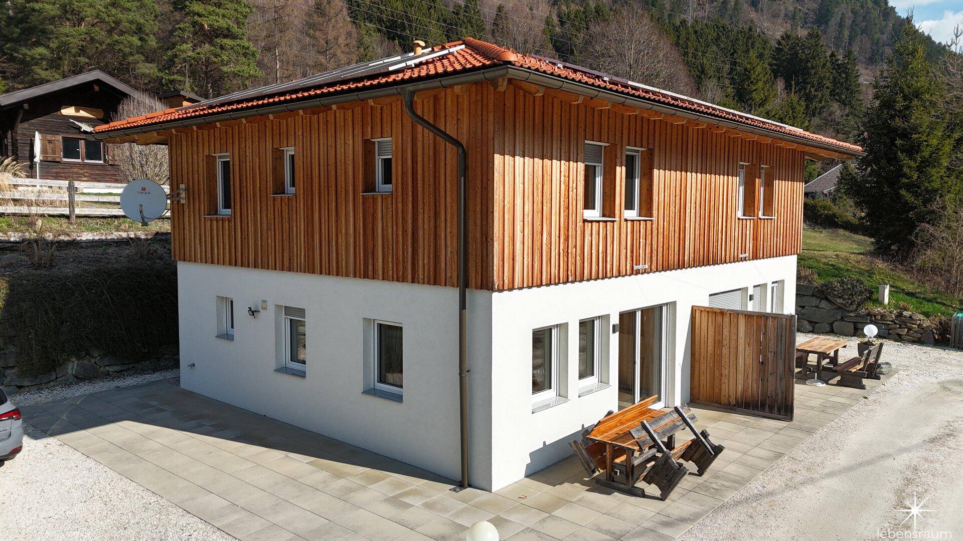 Seitenansicht des Hauses mit Holzfassade und weißem Putz, umgeben von grüner Landschaft.