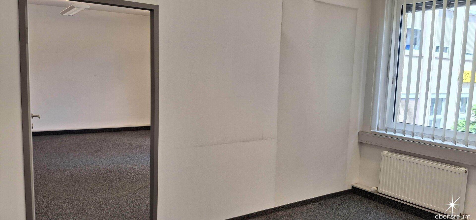 Helles Büro mit Teppichboden und einem Fenster, das viel natürliches Licht hereinlässt.