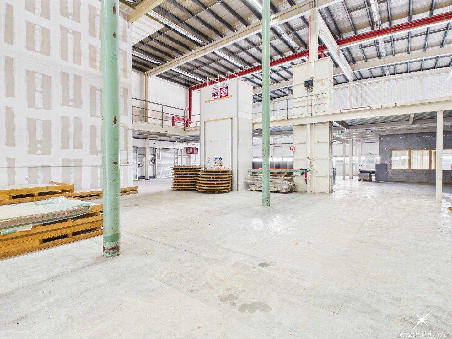 Weitläufige Industriehalle mit hohen Decken, Betonboden und einer erhöhten Plattform im Hintergrund.