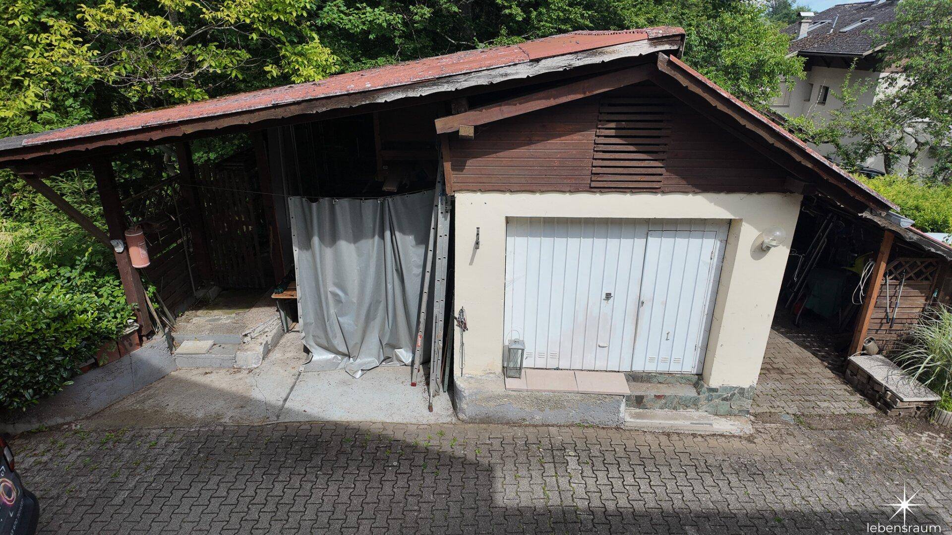 Separate Garage mit Holzverkleidung und angrenzendem Carport auf gepflastertem Vorplatz.