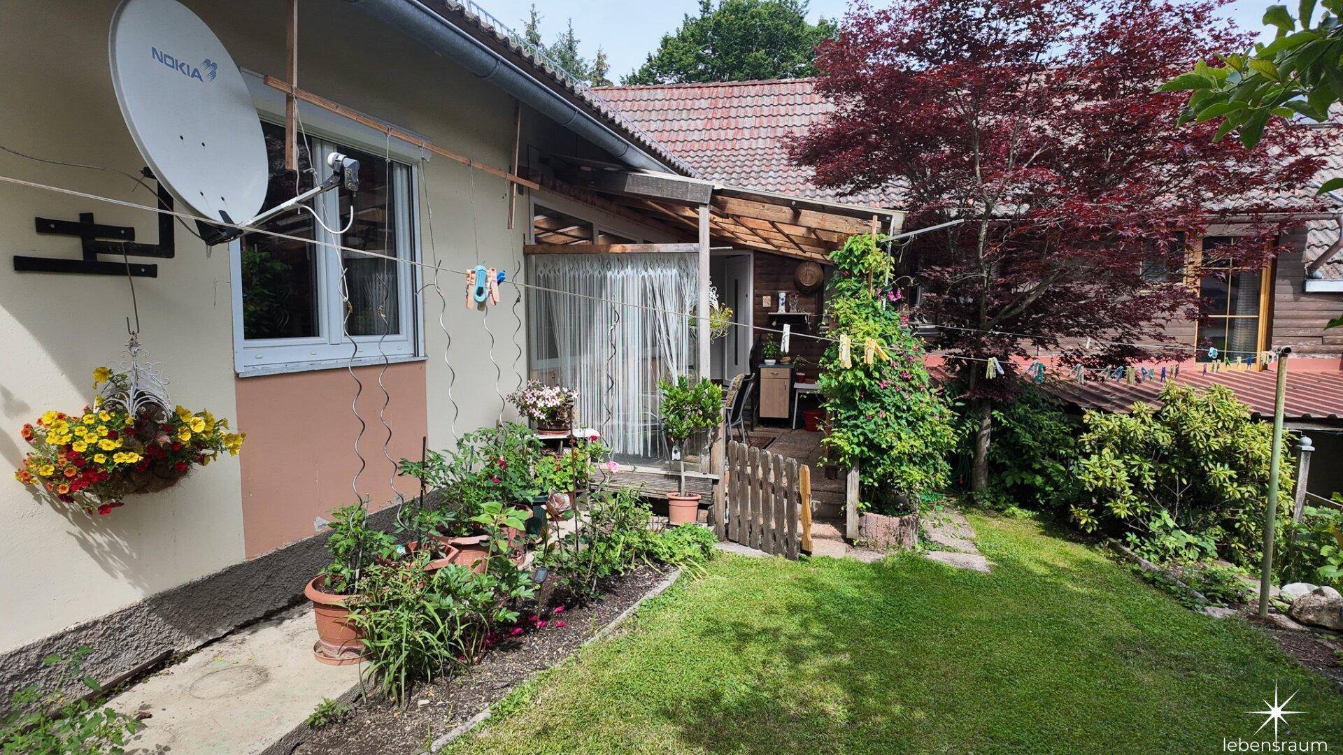 Seitlicher Gartenbereich mit Blumenbeeten, Rasenfläche und einem überdachten Sitzplatz am Haus.