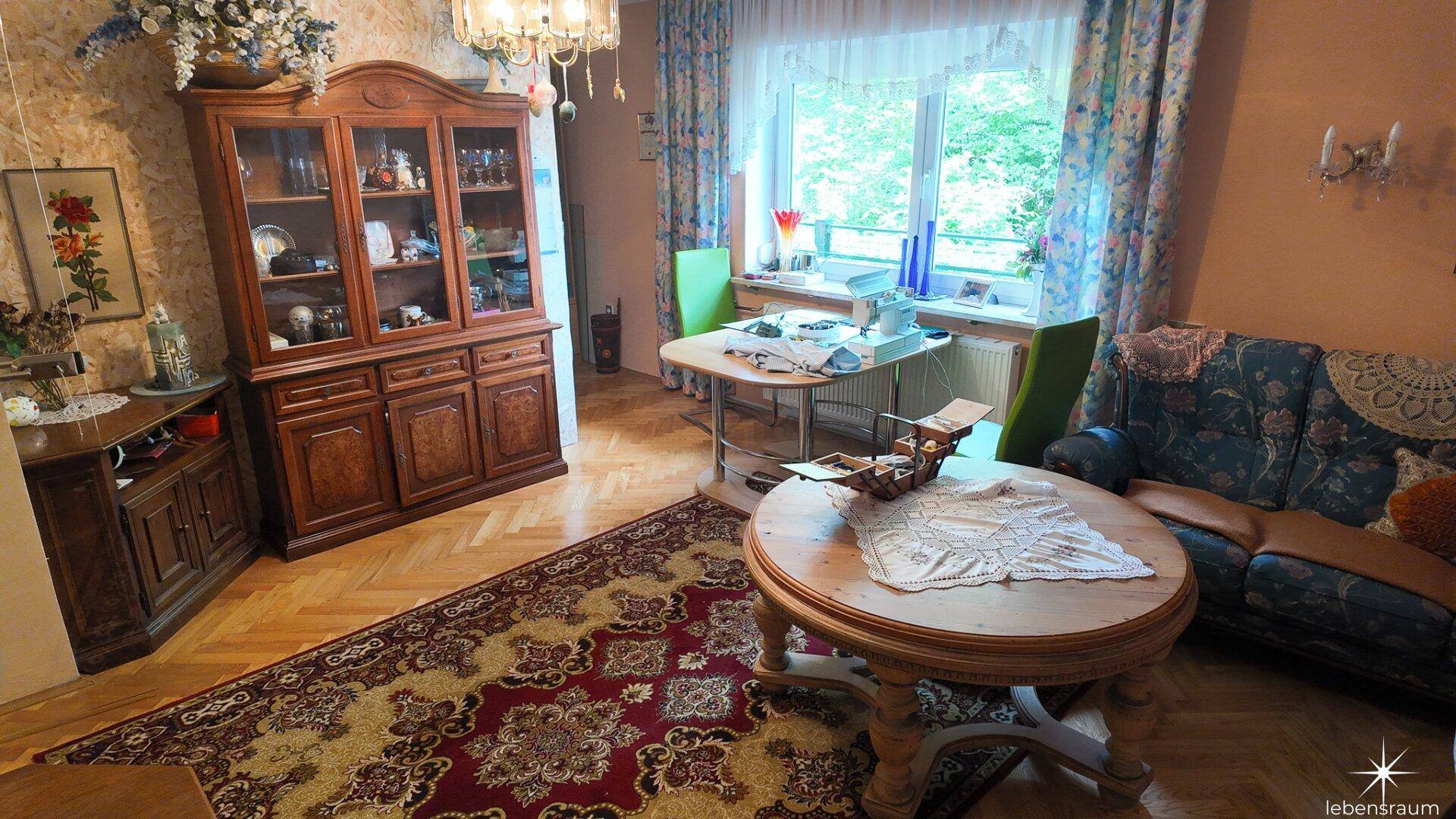 Wohnzimmer mit großem Fenster, Holzboden und traditionellen Möbeln, darunter ein Vitrinenschrank.