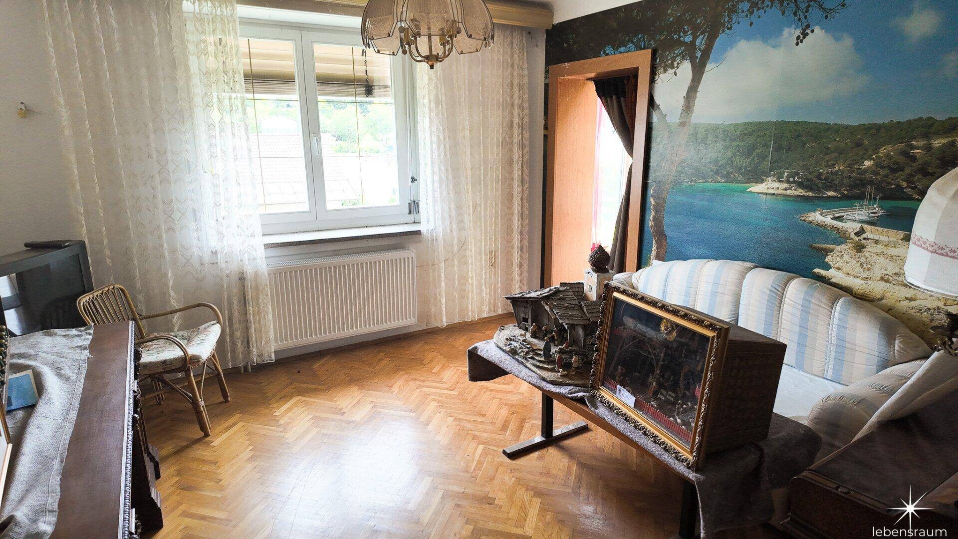 Wohnbereich mit Holzboden, großem Fenster und einer Wandtapete mit Meeresmotiv.