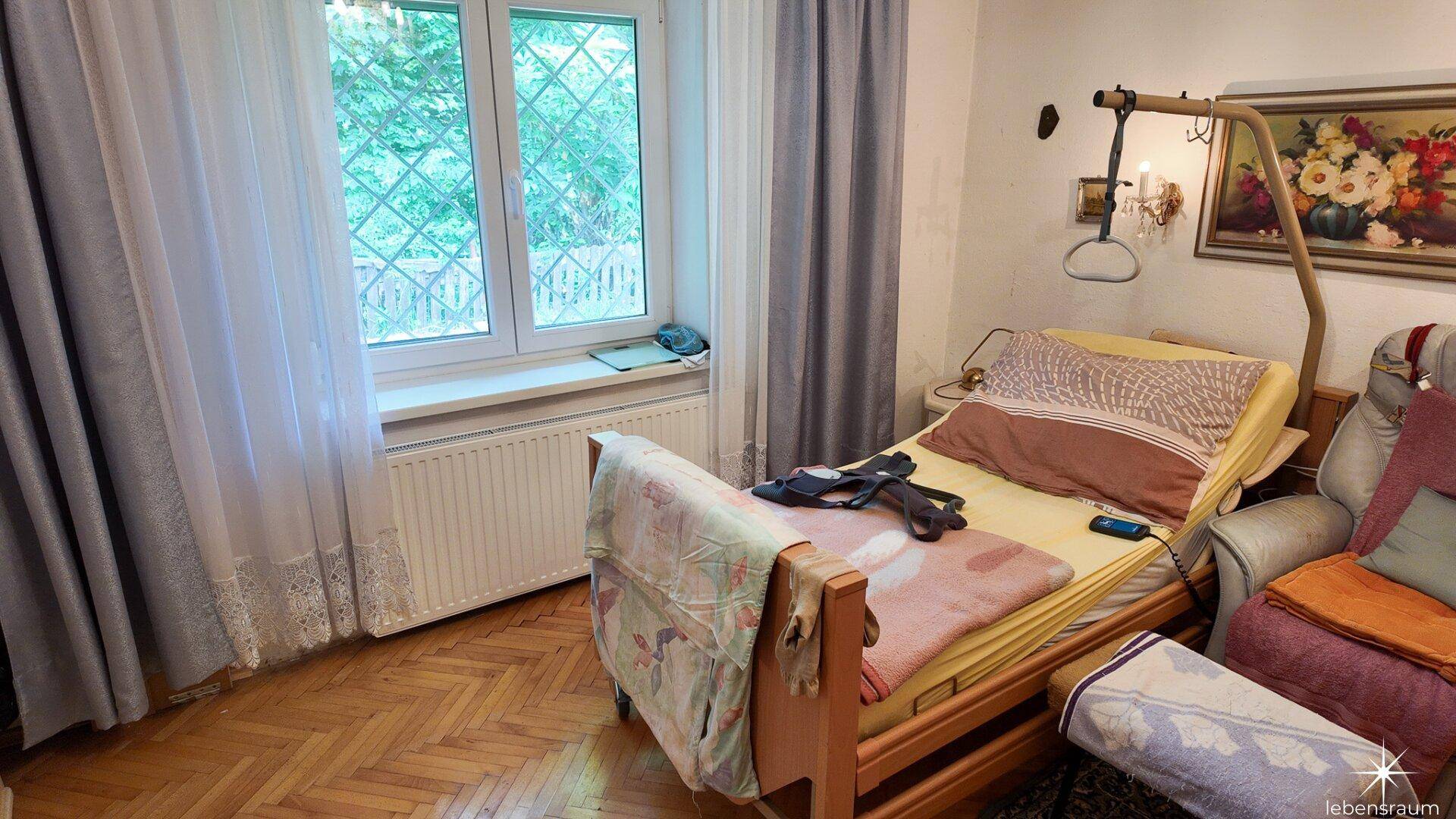 Schlafzimmer mit Pflegebett, Holzboden und Fenster mit Blick ins Grüne.