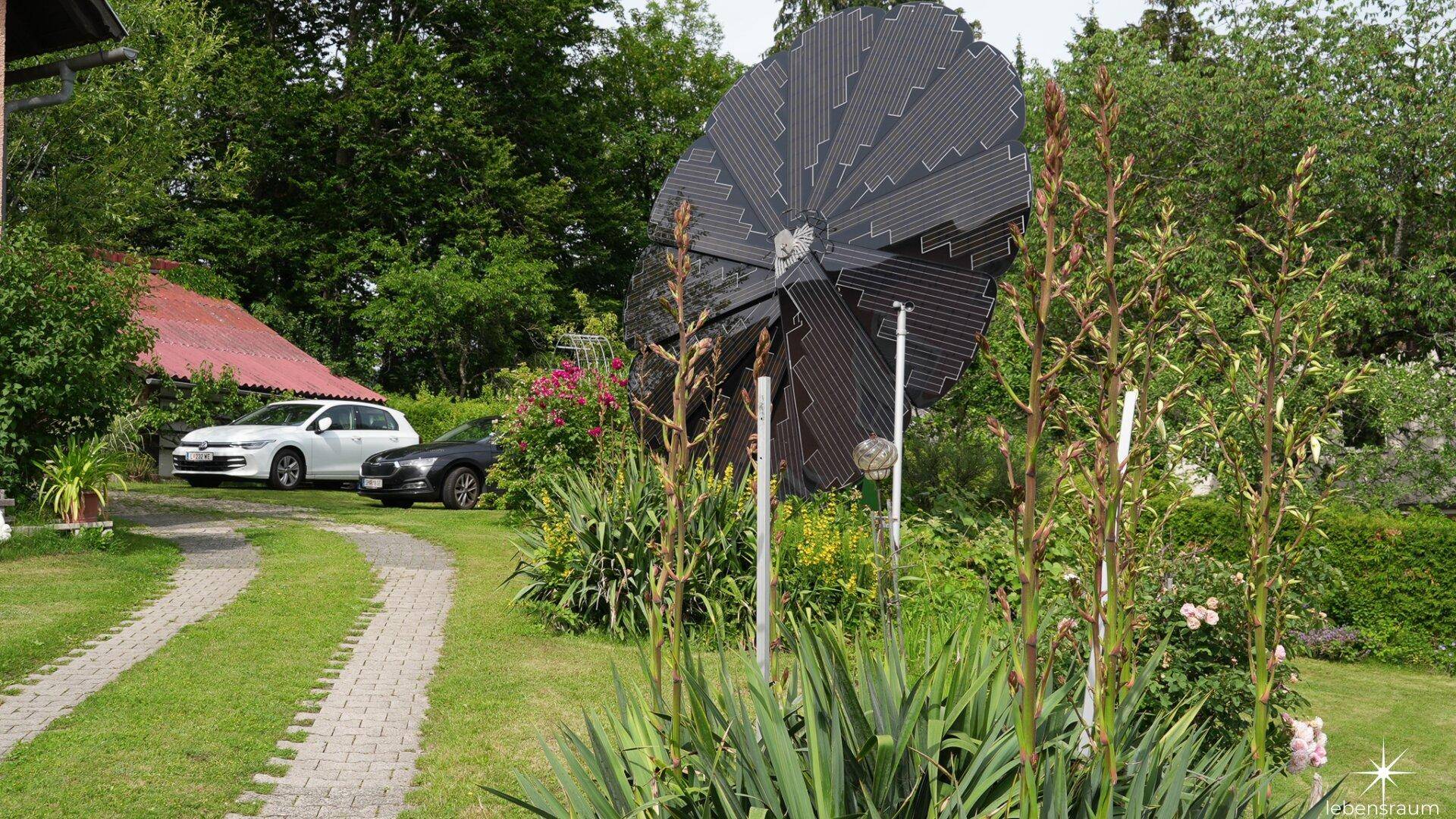 Garten mit dekorativer Solarblume, gepflasterter Auffahrt und üppiger Bepflanzung.
