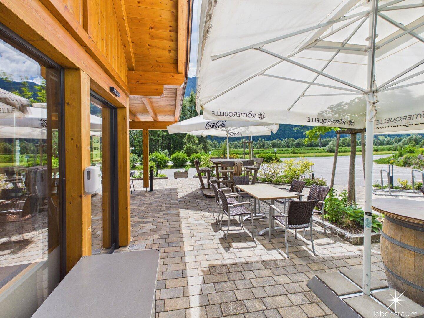 Sonniger Außenbereich mit gepflasterter Terrasse, Sonnenschirmen und Sitzgelegenheiten.