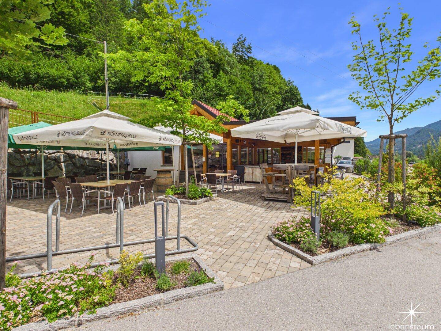 Weitläufiger Außenbereich mit gepflasterter Terrasse, Sonnenschirmen und Fahrradständern vor dem Gebäude.