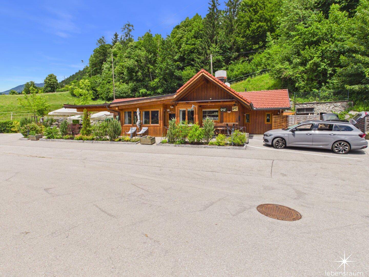 Holzverkleidetes Gebäude mit rotem Ziegeldach, umgeben von Parkplätzen und grüner Landschaft.