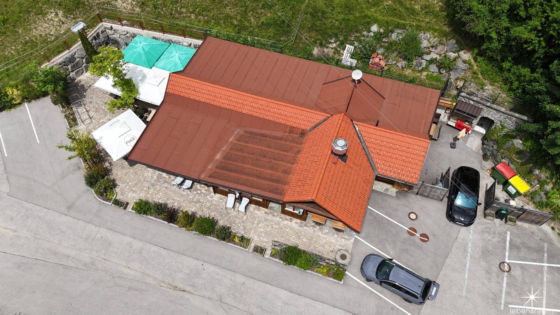Vogelperspektive eines Gebäudes mit rotem Ziegeldach, Terrasse und Parkplätzen.