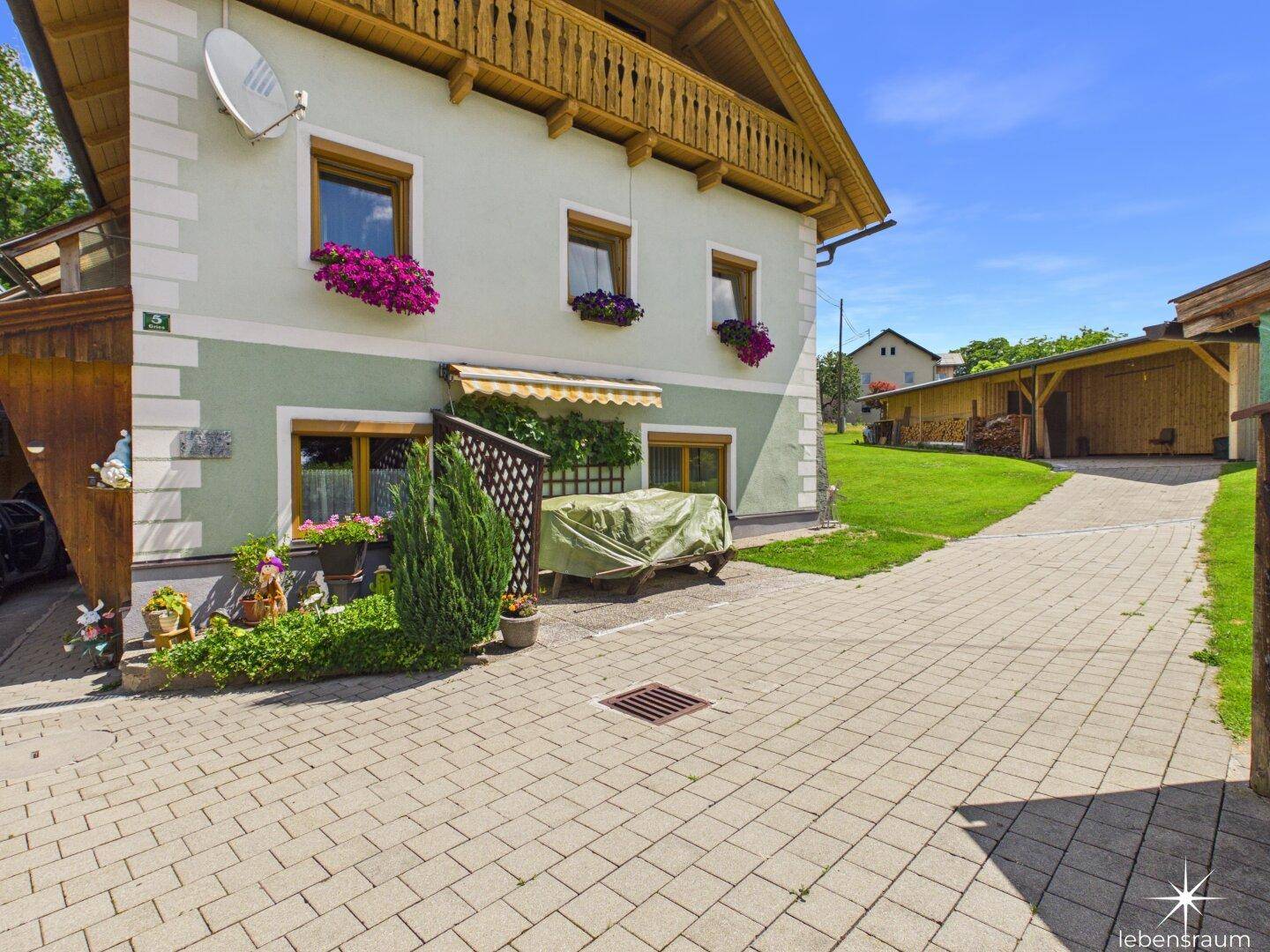 Traditionelles Haus mit gepflastertem Vorplatz, Blumenkästen und grüner Umgebung.