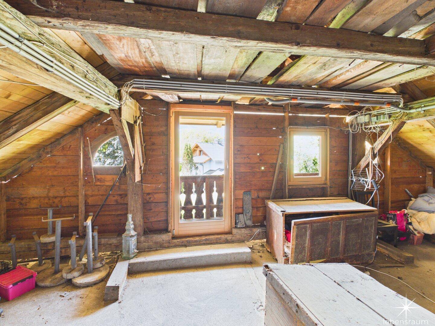 Unausgebauter Dachboden mit sichtbaren Holzbalken und Zugang zu einem kleinen Balkon.