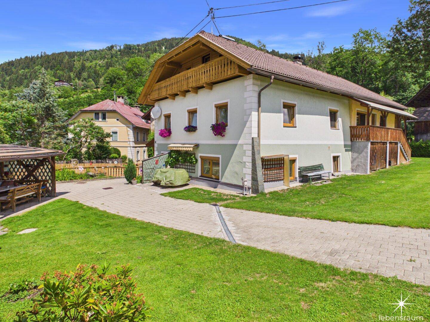 Charmantes Haus mit großem Garten, gepflasterter Zufahrt und Bergblick im Hintergrund.