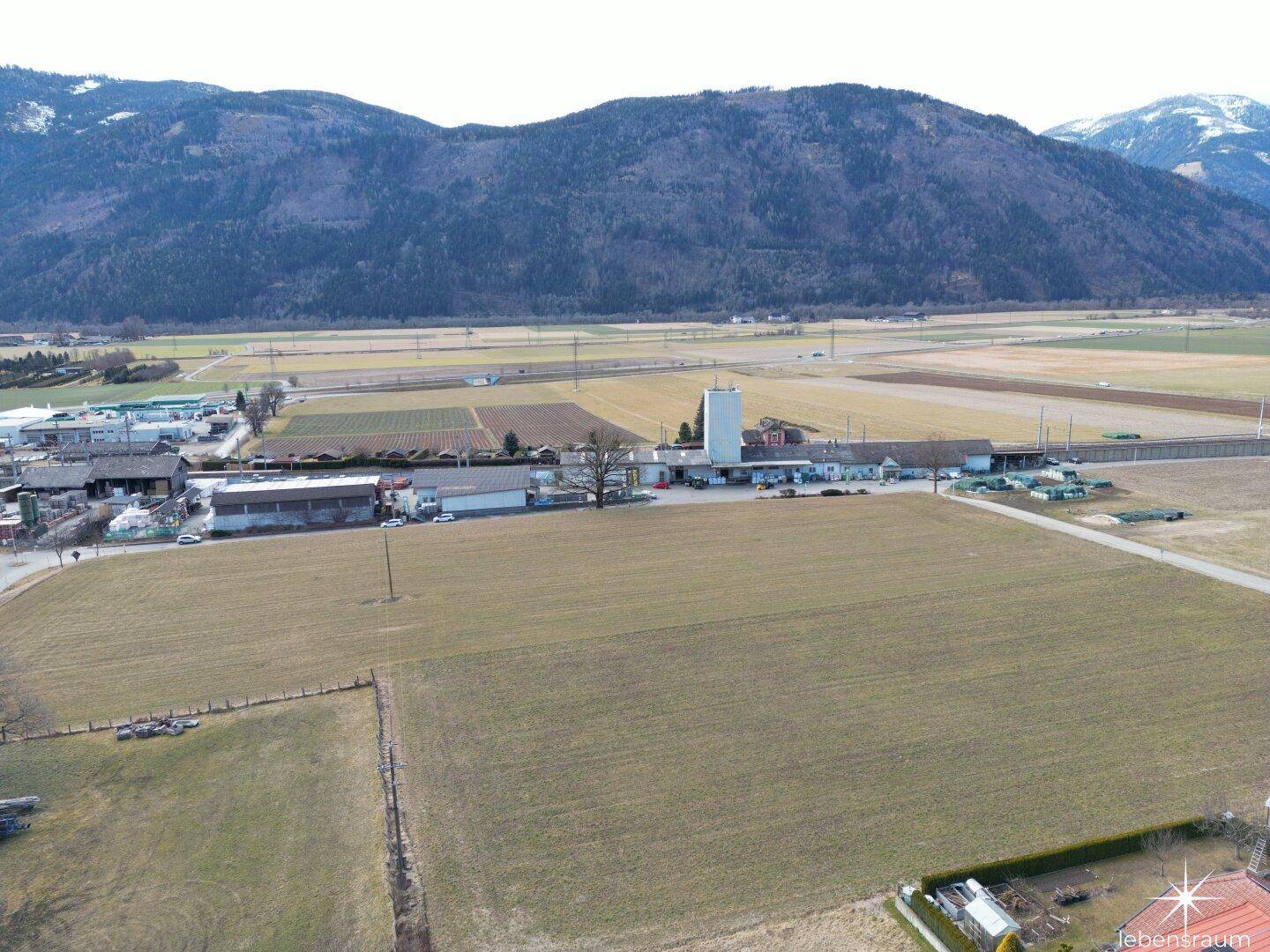 Weitläufige Luftaufnahme von Feldern, Industriegebäuden und einer beeindruckenden Bergkulisse.