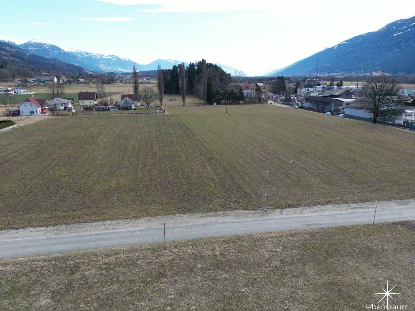 Luftaufnahme von weitläufigen Feldern, einer Landstraße und Häusern vor einer Bergkette.