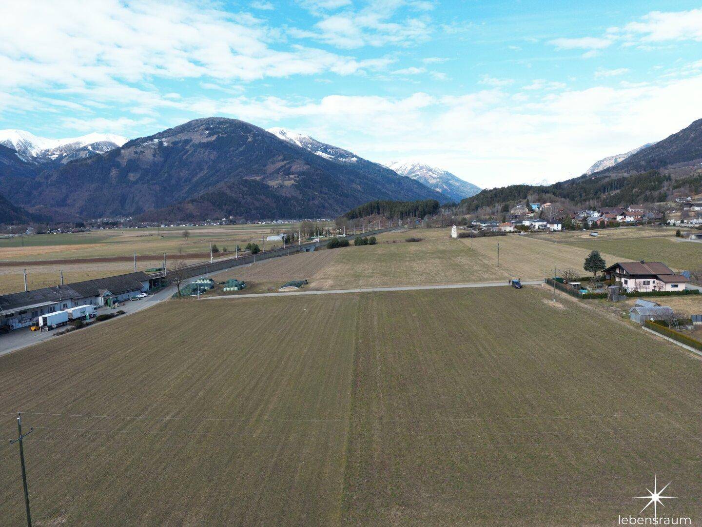 Weitwinkel-Luftaufnahme von grünen Feldern, vereinzelten Gebäuden und schneebedeckten Bergen.