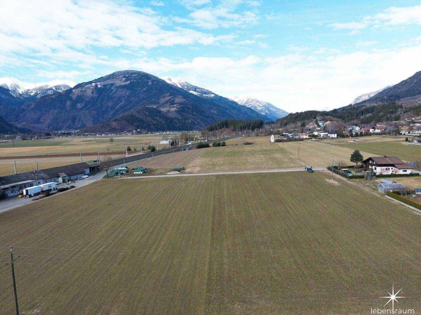 Weitwinkel-Luftaufnahme von grünen Feldern, vereinzelten Gebäuden und schneebedeckten Bergen.