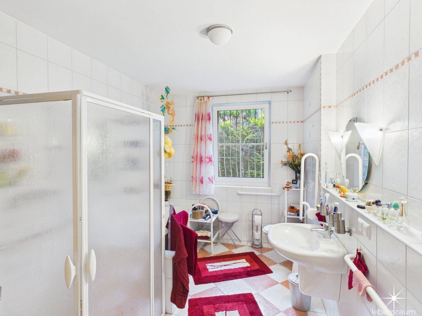 Badezimmer mit Duschkabine, Waschbecken und Fenster, dekoriert mit roten Akzenten.