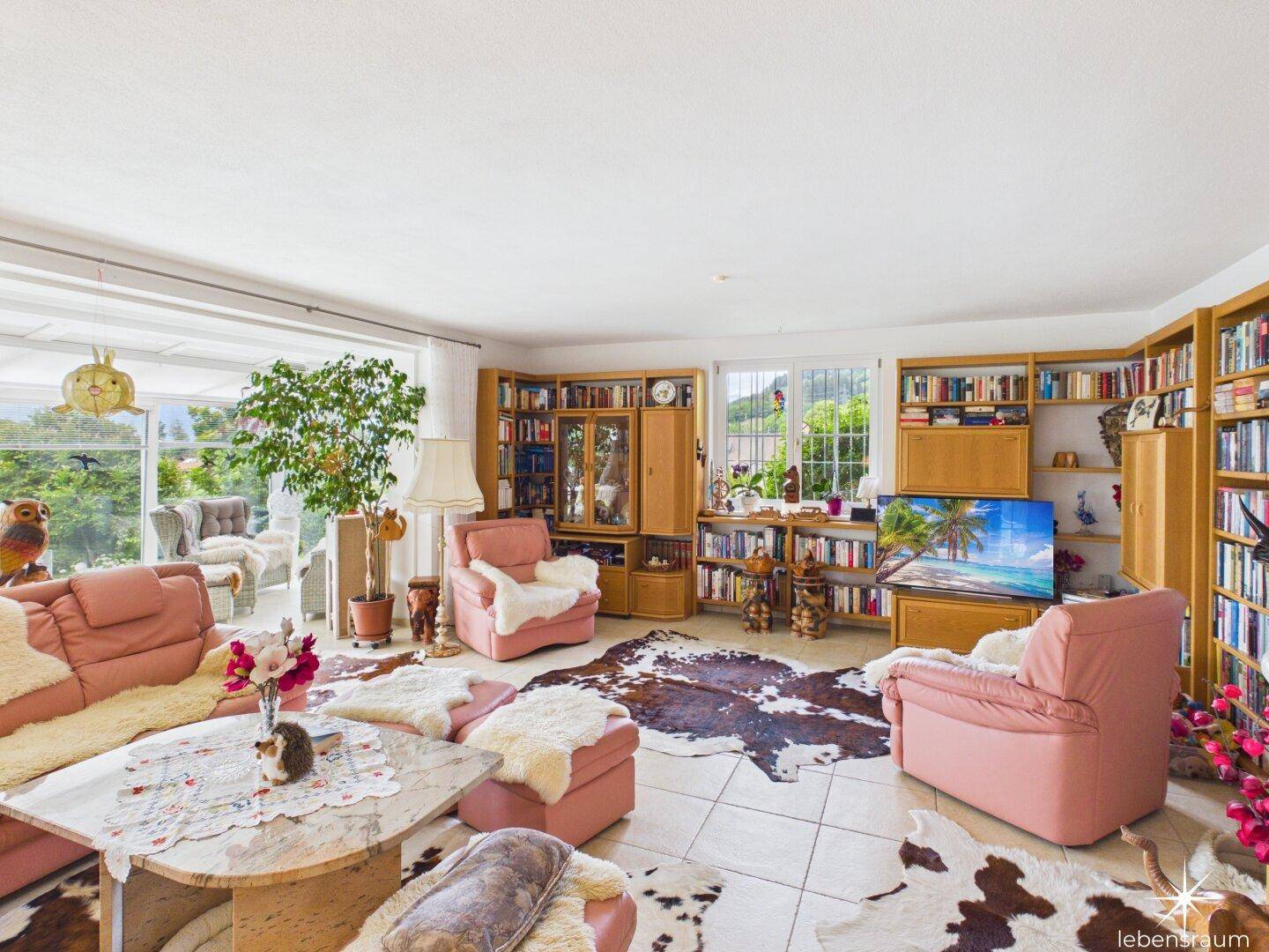 Helles Wohnzimmer mit umfangreichen Bücherregalen, rosa Sitzmöbeln und Zugang zum Wintergarten.