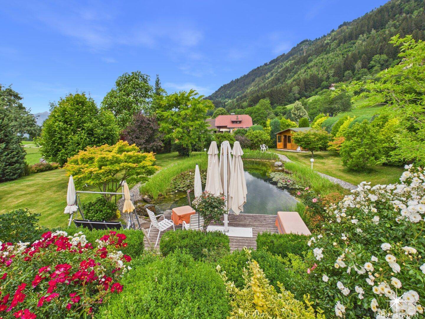 Malerischer Garten mit Teich, Sitzgelegenheiten und üppiger Bepflanzung vor Bergkulisse.