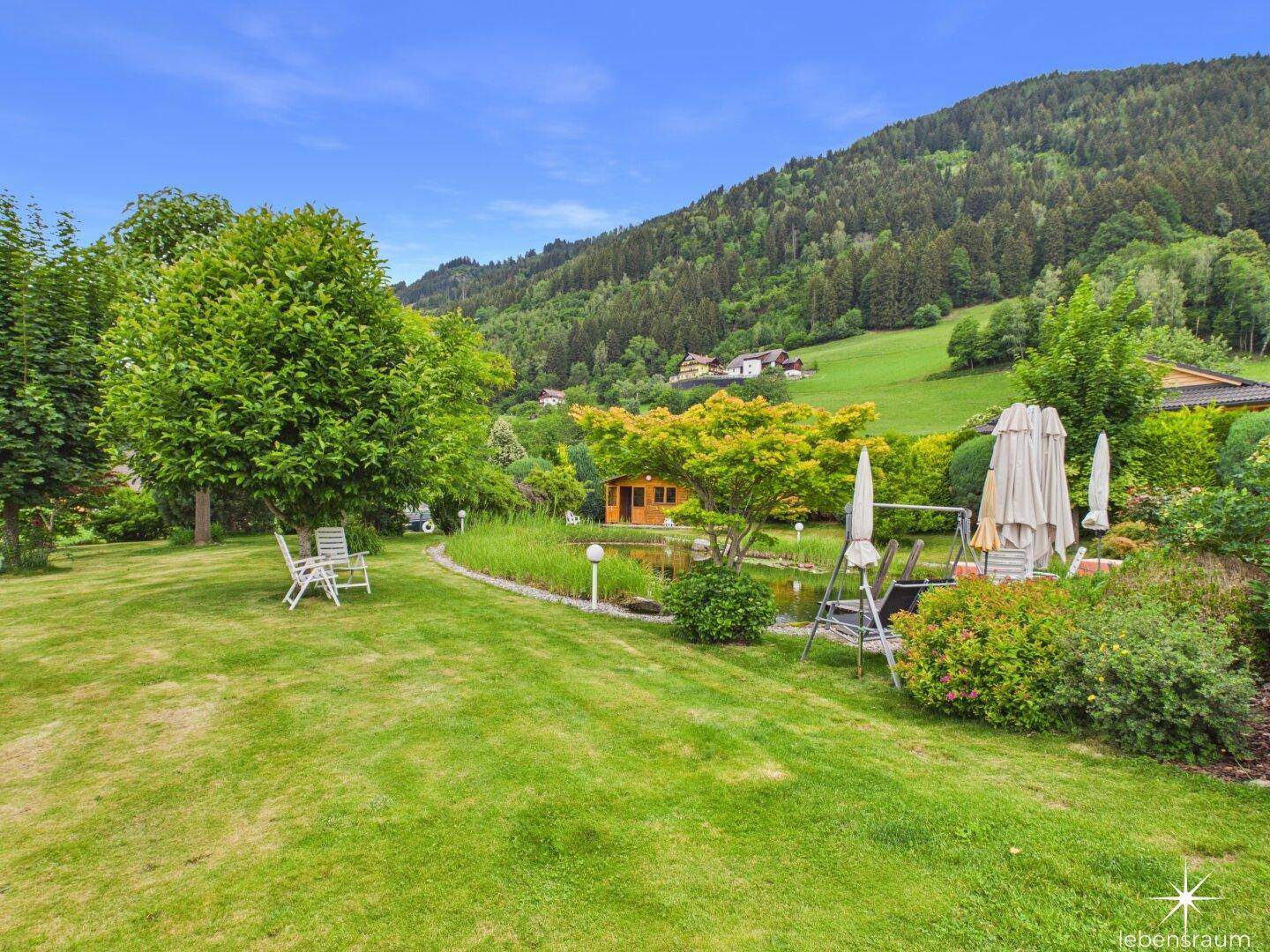 Weitläufiger Garten mit Rasen, Teich und Gartenhaus, umgeben von grünen Hügeln.