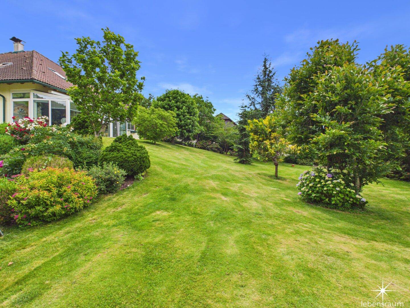Hügeliger Garten mit gepflegtem Rasen, Bäumen und Sträuchern, die das Haus umgeben.