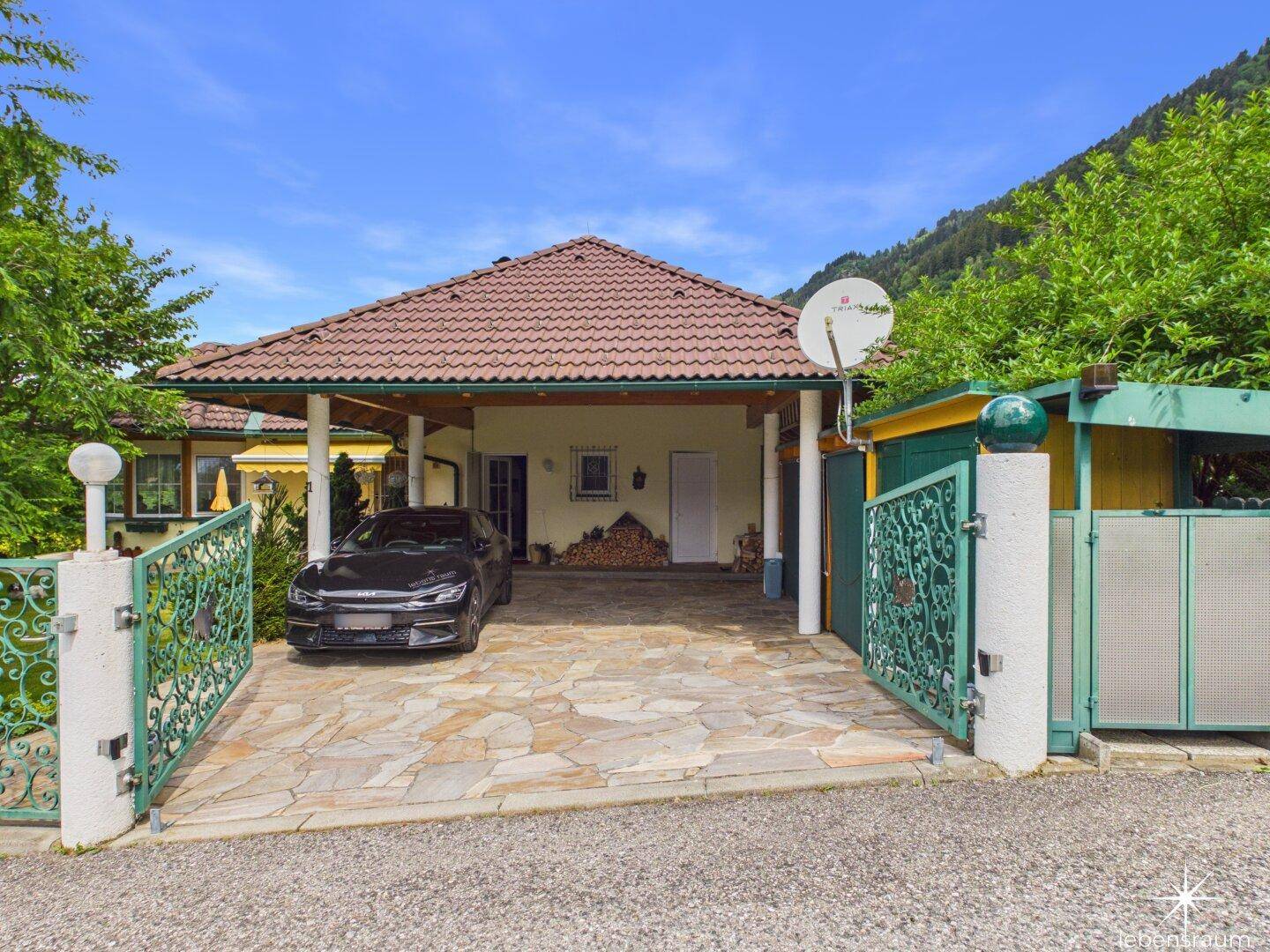 Einfahrt zum Haus mit Carport, gepflastertem Bereich und schmiedeeisernem Tor.