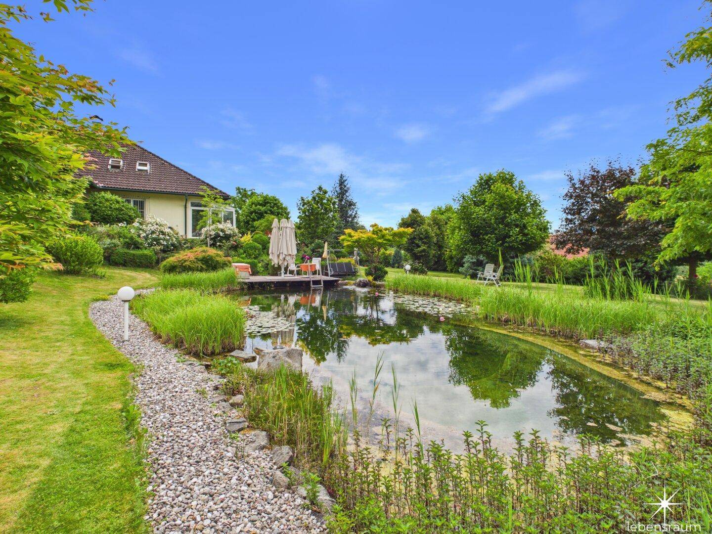 Großer Gartenteich mit Kiesweg, umgeben von üppiger Vegetation und dem Haupthaus.