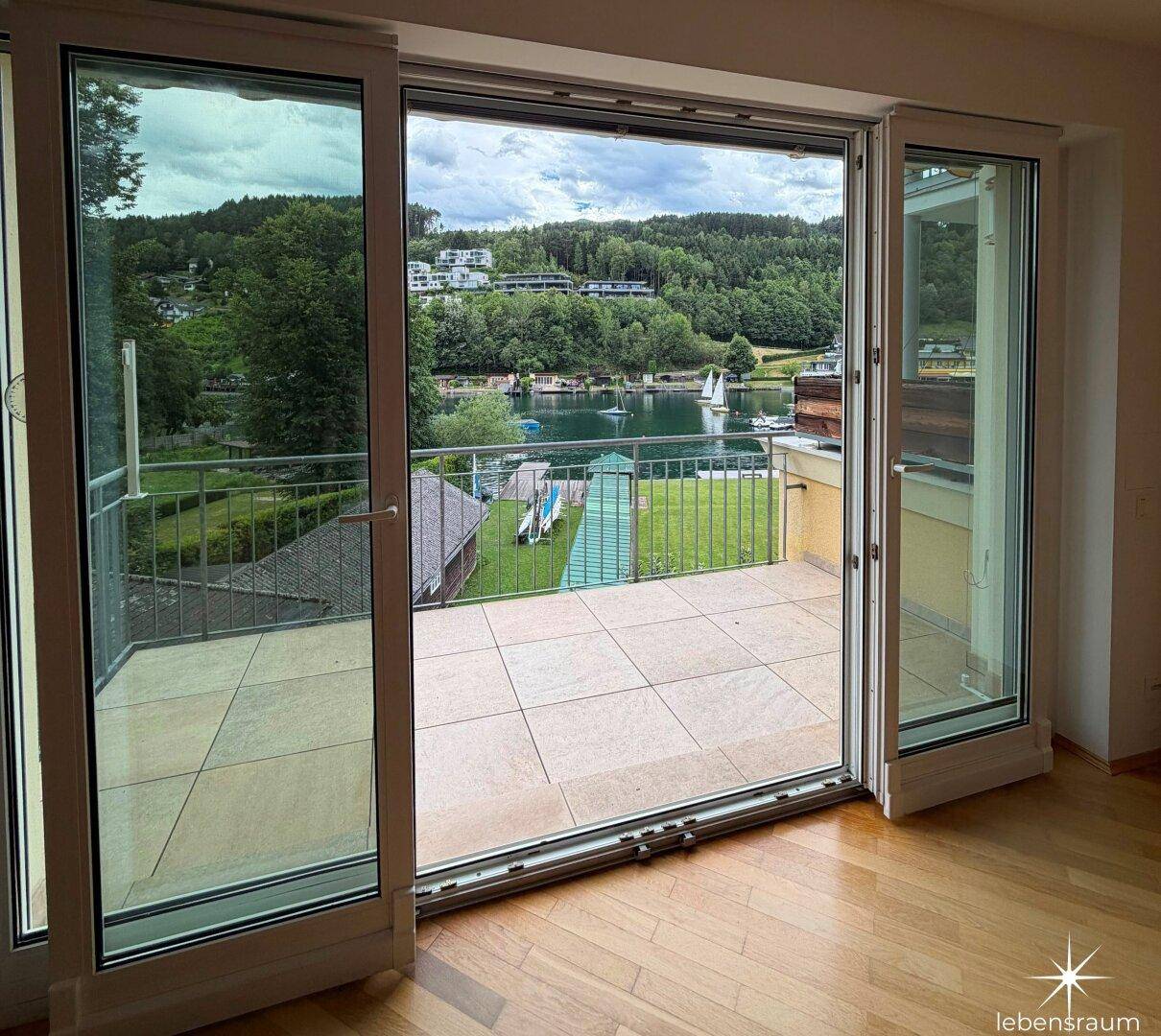 Blick aus dem Innenraum auf eine geflieste Terrasse mit weitem Seeblick und grüner Umgebung.